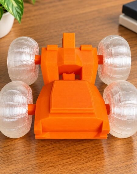 Vibrant orange 3D printed stunt car featuring translucent clear wheels on a wooden workstation