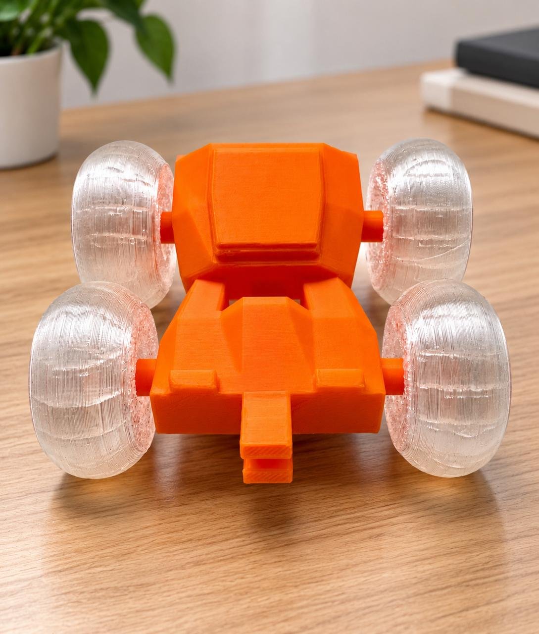 Vibrant orange 3D printed stunt car featuring translucent clear wheels on a wooden workstation